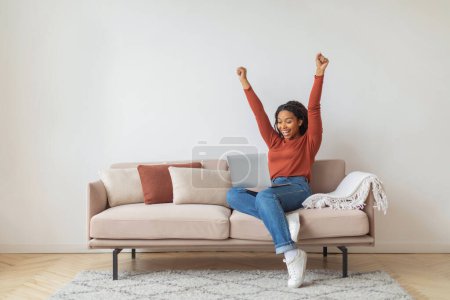 Foto de Mujer negra alegre levantando sus brazos en la victoria mientras está sentada con el ordenador portátil en el sofá, mujer afroamericana celebrando el éxito - Imagen libre de derechos