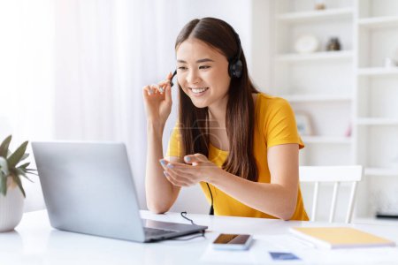 Enganchado joven asiática mujer usando auriculares y sonriendo en la pantalla del ordenador portátil, hablando en una videollamada en casa oficina