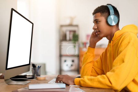 A focused man in a yellow hoodie is intently working on a project, using a large desktop computer in a cozy home setting