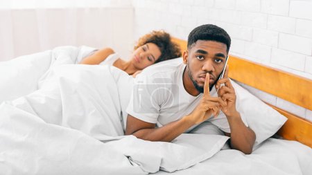 African American man looks worried during a phone call as his partner naps, reflecting relationship and communication themes