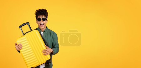 Foto de Un chico afro americano amigable y alegre en gafas de sol abraza una maleta de color amarillo brillante, mostrando felicidad y afecto por viajar sobre un fondo amarillo - Imagen libre de derechos
