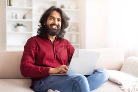 A relaxed Indian guy with a friendly demeanor working on a laptop, comfortably seated on a couch in a bright room