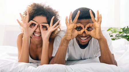Téléchargez les photos : Joyeux couple afro-américain heureux amoureux s'amuser au lit, tenant les doigts près des yeux - en image libre de droit
