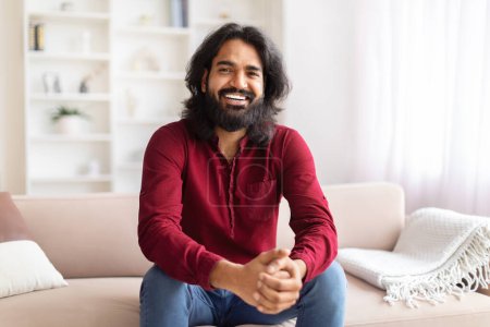 Téléchargez les photos : Homme indien joyeux aux cheveux longs et à la barbe confortablement assis sur un canapé dans un salon lumineux et confortable - en image libre de droit