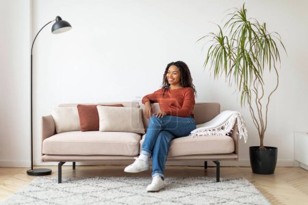 Téléchargez les photos : Heureuse femme noire assise sur un canapé, entourée d'un décor minimaliste, profitant d'un moment paisible. - en image libre de droit