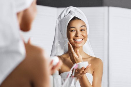 Foto de Sonriente afro-americana aplicando crema en su cara en el baño, reflejándose en el espejo. - Imagen libre de derechos