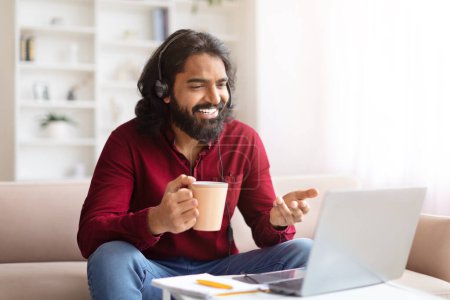 Bearded Indian man with headphones on laughing during a vide call on a laptop, have online meeting, drinking coffee