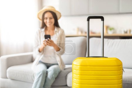 Yellow suitcase in focus with a blurred background where a woman using a smartphone is visible, booking taxi