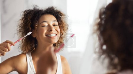 Téléchargez les photos : Jeune femme afro souriante avec brosse à dents nettoyant les dents dans la salle de bain. Concept de dents saines - en image libre de droit