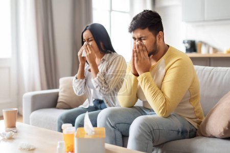 Indian man and woman are blowing their noses while sitting apart on a sofa, tissues and a mug visible.