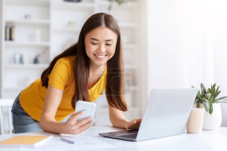 Téléchargez les photos : Souriant asiatique femme en haut jaune en utilisant un téléphone portable et un ordinateur portable simultanément à son bureau, travaillant à partir de la maison - en image libre de droit