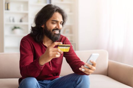 Téléchargez les photos : Homme indien joyeux tenant une carte de crédit et regardant le téléphone mobile pour les achats en ligne, intérieur de la maison - en image libre de droit