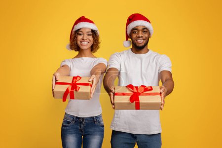 Téléchargez les photos : Un jeune couple afro-américain joyeux portant des chapeaux de Père Noël présente des cadeaux de Noël sur un fond jaune vibrant - en image libre de droit