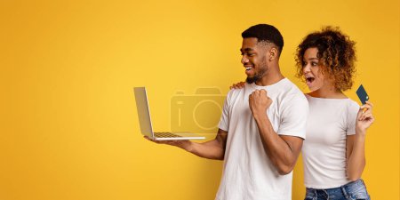 Foto de Una alegre pareja afroamericana joven sosteniendo una computadora portátil y una tarjeta de crédito, expresando emoción por las compras en línea sobre un fondo amarillo - Imagen libre de derechos