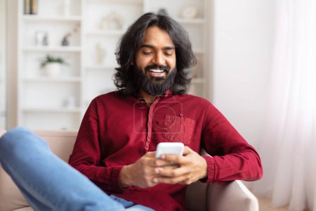 Téléchargez les photos : Homme indien assis sur un canapé et utilisant son smartphone, regarder le contenu dans un cadre chaleureux et accueillant à la maison - en image libre de droit