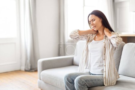 Photo for A woman on a couch appears uncomfortable, with her hands placed on her neck, indicating soreness or stress - Royalty Free Image