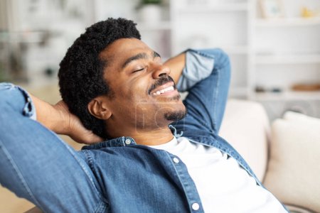 Foto de Hombre negro está descansando en un sofá en casa con las manos colocadas detrás de la cabeza en una posición relajada, disparo de primer plano - Imagen libre de derechos