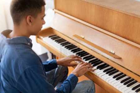 Photo for Rear view of dedicated teenager guy wearing denim shirt, deeply concentrated while practicing on the wooden piano keys in sunny room - Royalty Free Image
