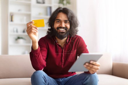 Hombre indio alegre mostrando tarjeta de crédito, usando tableta digital, posiblemente haciendo una compra desde casa