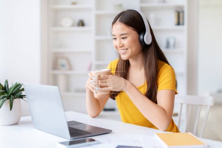 Téléchargez les photos : Sourire femme asiatique dans un haut jaune profiter du café lors d'un appel vidéo à son espace de travail, en utilisant un ordinateur portable et écouteurs - en image libre de droit