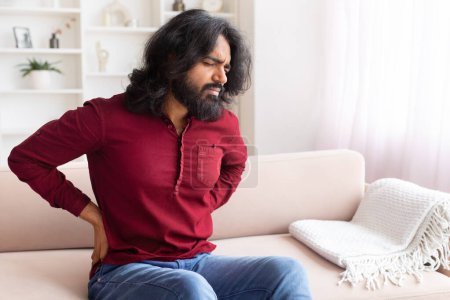 Chico indio que experimenta dolor de espalda, ligeramente inclinado hacia adelante, expresando malestar mientras está sentado en un sofá elegante