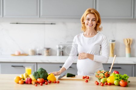 Téléchargez les photos : Femme enceinte souriante préparant une salade de légumes dans une cuisine moderne et bien équipée - en image libre de droit