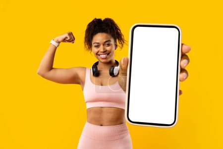 Energetic fit mujer afroamericana con auriculares presenta alegremente un teléfono inteligente con una pantalla en blanco sobre un fondo amarillo