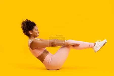Téléchargez les photos : Jeune femme afro-américaine active en tenue de sport rose tient une pose de bateau, technique Pilates sur fond jaune - en image libre de droit