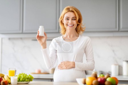Téléchargez les photos : Joyeux attente mère tenant une bouteille de vitamines prénatales dans une cuisine bien approvisionnée - en image libre de droit
