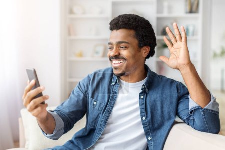 Téléchargez les photos : Un homme noir joyeux agitant à l'écran tout en tenant un smartphone, s'engageant dans un appel vidéo à la maison - en image libre de droit