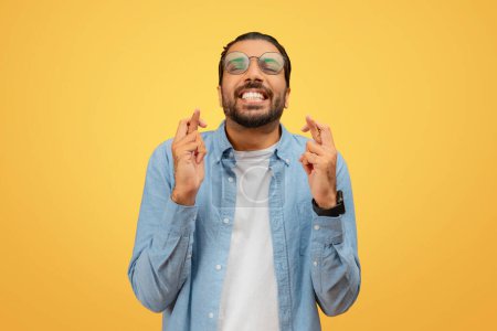 Indian man in a relaxed blue shirt has his eyes closed, crossing his fingers in hope on a yellow background