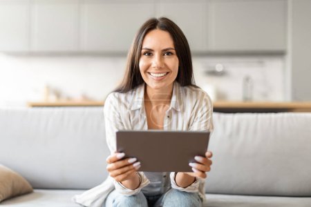 Eine fröhliche Frau, die bequem mit einem digitalen Tablet sitzt und im häuslichen Umfeld in die Kamera blickt