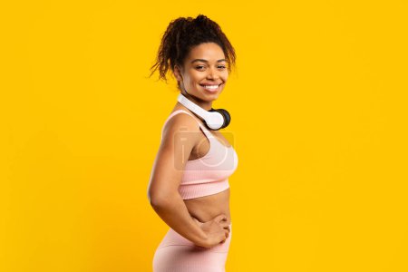Femme afro-américaine souriante et confiante dans des poses de vêtements de sport avec des écouteurs autour du cou, sur fond jaune joyeux