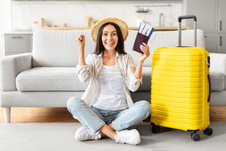 Délicieuse femme tenant un passeport et une carte d'embarquement, avec une valise jaune, célébrant les prochains voyages