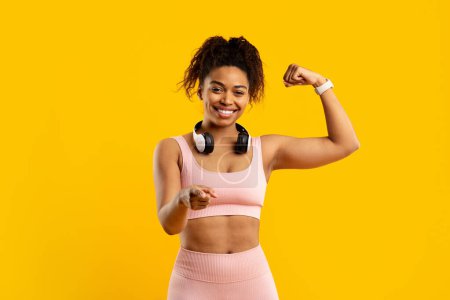 Smiling african american woman confidently shows her muscles with fists up in a power stance, dressed in gym attire, against yellow