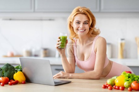 Foto de Ajuste mujer rubia disfrutando de un batido verde mientras trabaja en un ordenador portátil en una cocina organizada - Imagen libre de derechos