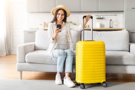 Cheerful woman with a straw hat checks her smartphone beside a yellow suitcase, in a comfortable home