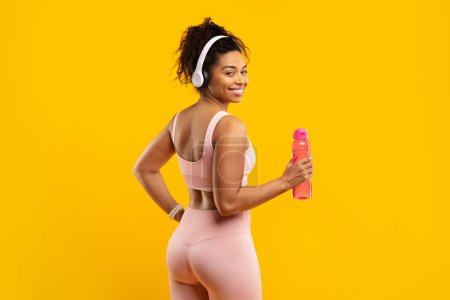Téléchargez les photos : Fit afro-américaine en tenue rose sourit tout en tenant une bouteille d'eau et le port d'écouteurs, prêt pour une séance de gymnastique - en image libre de droit