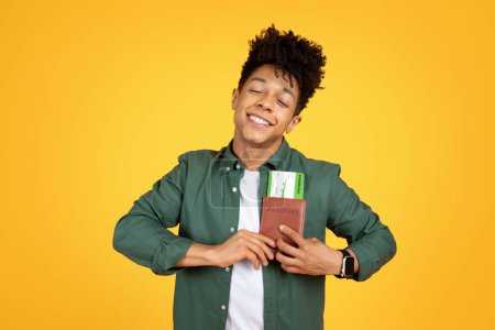 Téléchargez les photos : Un homme afro-américain satisfait tient doucement un passeport, évoquant des sentiments de satisfaction et de désir de voyager sur un fond jaune - en image libre de droit