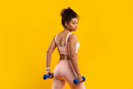 Photo for A strong african american woman confidently lifts blue dumbbells during her fitness session, showcasing strength on a yellow backdrop - Royalty Free Image
