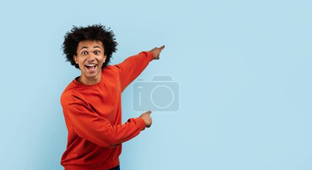 Téléchargez les photos : Un homme noir enthousiaste dans un pull rouge pointe loin avec un regard surpris sur un fond bleu - en image libre de droit