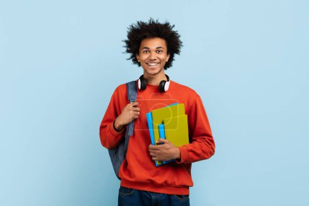 Un joven estudiante afroamericano sonriente con auriculares alrededor del cuello lleva libros sobre fondo azul