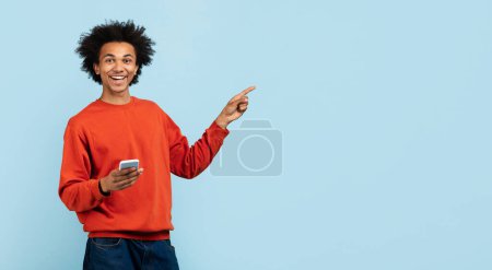 Téléchargez les photos : Heureux jeune homme afro-américain tenant un smartphone, pointant joyeusement vers le côté sur fond bleu - en image libre de droit