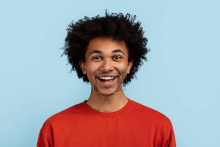 Un jeune homme noir joyeux aux cheveux bouclés portant un pull rouge sourit sur un fond bleu