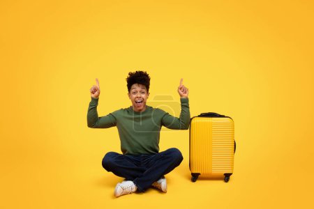 Photo for An excited young black man with raised hands sitting next to a yellow rolling suitcase on a yellow background, copy space - Royalty Free Image