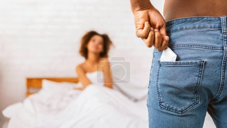 Photo for Passion, desire games and sexual health. Black man taking condom from jeans, naked woman waiting in bed, free space - Royalty Free Image
