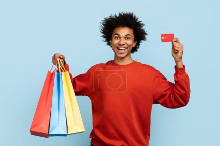 Exuberante hombre afroamericano sosteniendo coloridas bolsas de compras y una tarjeta de crédito sobre fondo azul