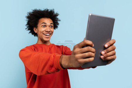 A delighted african american man takes a selfie with a digital tablet, expressing happy astonishment