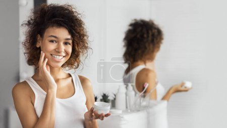 Joyeux afro fille appliquant de la crème sur son visage, debout dans la salle de bain, réfléchissant dans le miroir.