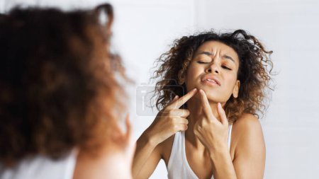 Photo for Afro girl frowning squeezing pimple on her face in mirror. Do not squeez pimples concept, copy space - Royalty Free Image